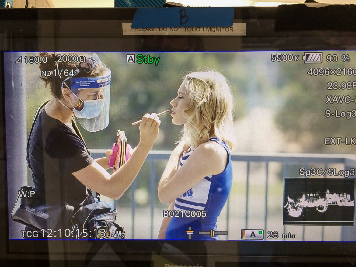 thumbnail_Makeup re-touching on Cheerleader Hit Squad September 2020 Makeup artist in mask and face shield retouching makeup of blonde actress in blue and white cheerleading uniform.